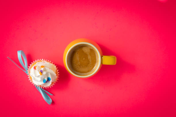 Small yellow cup filled with double espresso shots in the middle of bright pink background. White frosting cupcake with light blue bow tie and colorful sprinkles placed next to the coffee.