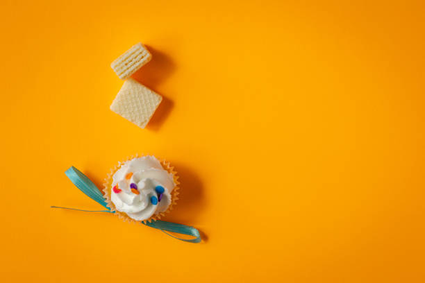 Top down view of white icing cupcake with colorful sprinkles and two party size wafers placed on bright yellow background