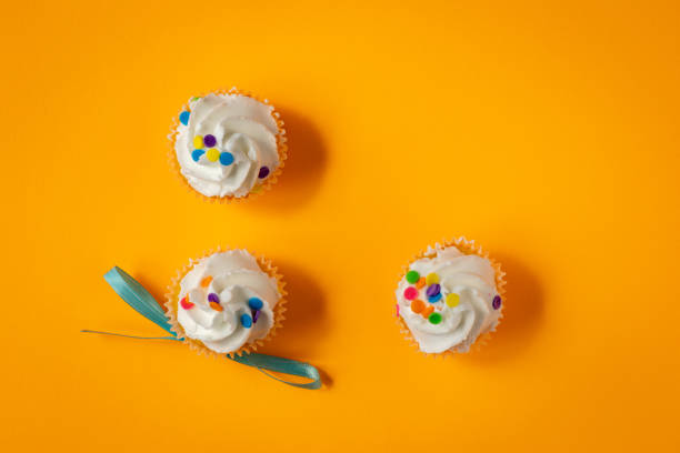 Top down view of three white frosting cupcakes with multicolored sprinkles on bright orange background.