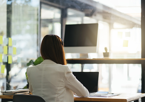 Beautiful Businesswoman Taking Place at Her Working Desk in the Office with Cityscape Window View. Successful Woman Wearing Formal Wear Dress Working on a Laptop. Descending Back View Shot.