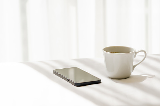 Selective focus of a cup of black coffee and smartphone put on white bed sheet with shadow of curtain in the morning. Relaxing at home concept.