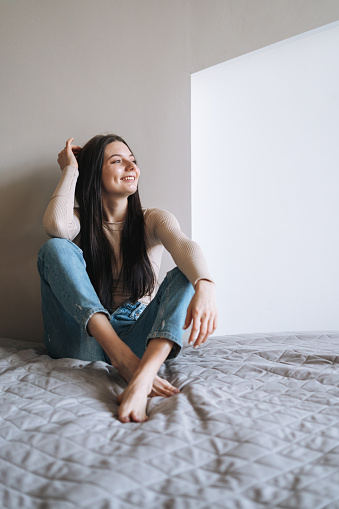 Young smiling woman teenager girl with dark long hair in jeans sitting on bed in room