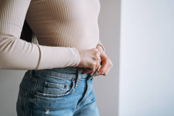 Crop photo of Young smiling woman teenager girl with dark long hair in jeans sitting on bed at the home Crop photo of Young smiling woman teenager girl with dark long hair in jeans sitting on bed at home woman hand in pants stock pictures, royalty-free photos & images