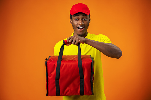 Smiling black male courier wearing uniform holding box in orange studio, close up