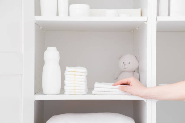 young adult mother hand taking white towel from inside wardrobe. bottle of talcum powder, stack of new nappies and teddy bear on shelf. preparing for baby diaper changing. hygiene goods. closeup. - baby goods imagens e fotografias de stock