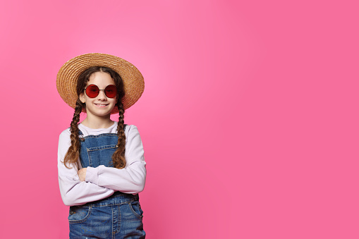 Cute little girl in straw hat and red sunglasses, looking at camera with crossed hands, isolated pink background. Space for text.