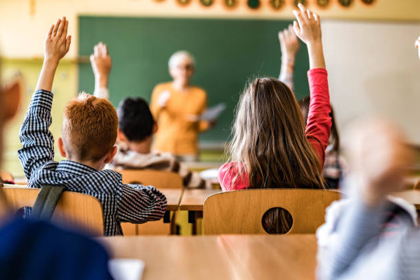 istock Rear view of elementary students raising their hands on a class. 1386826077