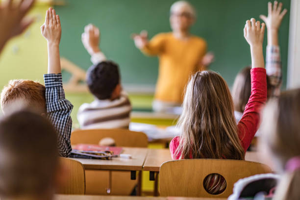 istock Back view of students raising their hands to answer the teacher's question on a class. 1386826148