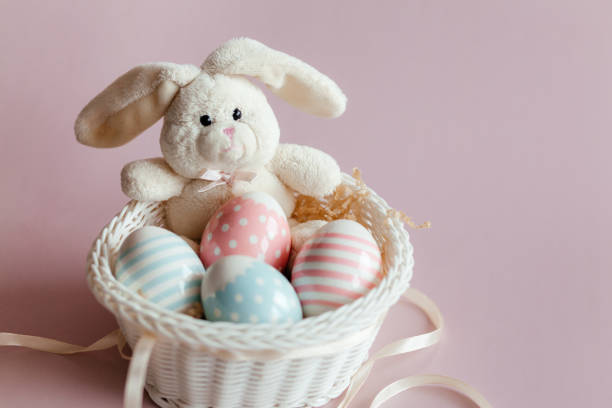 Easter eggs in pastel colors with a pattern and a white toy rabbit in a basket on a pink background