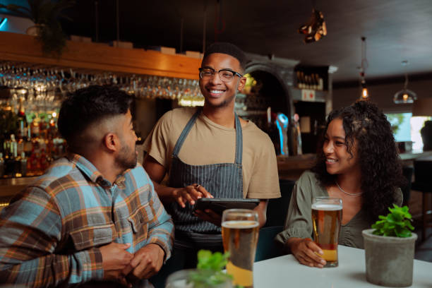 African American male waiter serving couple and friends at the restaurant African American male waiter serving couple in restaurant using digital tablet. High quality photo busy restaurant tables stock pictures, royalty-free photos & images