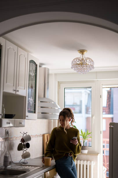 istock Woman standing in the kitchen and drinking cup of tea while using smartphone 1386837834