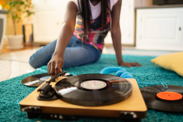 A record player with a long playing record on it as an unrecognizable girl is reaching out to place the needle onto the record, more vinyl is lying on the rug around the stereo