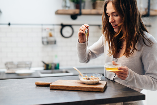 Young woman in the kitchen adds honey to granola.