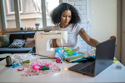 A young businesswoman is checking details on her laptop, while sewing items with colorful materials, sitting at her dining table at home