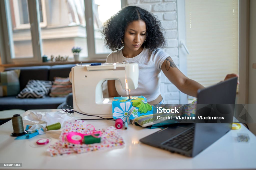 Young seamstress businesswoman using laptop and sewing machine A young businesswoman is checking details on her laptop, while sewing items with colorful materials, sitting at her dining table at home Sewing Machine Stock Photo