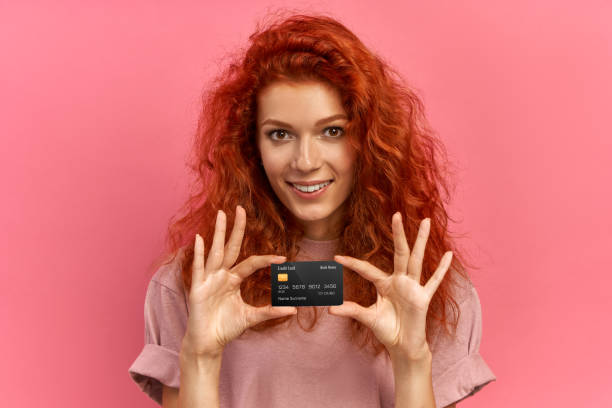 tiempo de compras. joven mujer sonriente de jengibre con apariencia atractiva, demostrando tarjeta de plástico, alegrada de recibir el salario yendo a comprar ropa nueva en la tienda de ropa, aislada sobre fondo rosa - greeting card holding women credit card fotografías e imágenes de stock