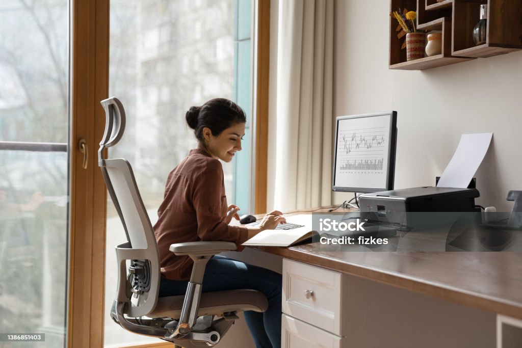 Concentrated indian woman working in home office. Concentrated young indian ethnicity woman sitting in comfortable adjustable ergonomic armchair with lumbar support, studying or working on computer in modern home office. distant workday concept. Ergonomics Stock Photo