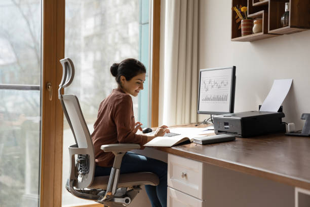 Concentrated indian woman working in home office. Concentrated young indian ethnicity woman sitting in comfortable adjustable ergonomic armchair with lumbar support, studying or working on computer in modern home office. distant workday concept. ergonomics stock pictures, royalty-free photos & images