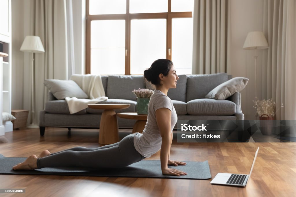 Happy young indian woman stretching muscles, enjoying online yoga class. Full length flexible happy young indian woman stretching muscles doing Bhujangasana Cobra pose, enjoying yoga online training watching video lecture distant online workout on computer at home. Spine - Body Part Stock Photo