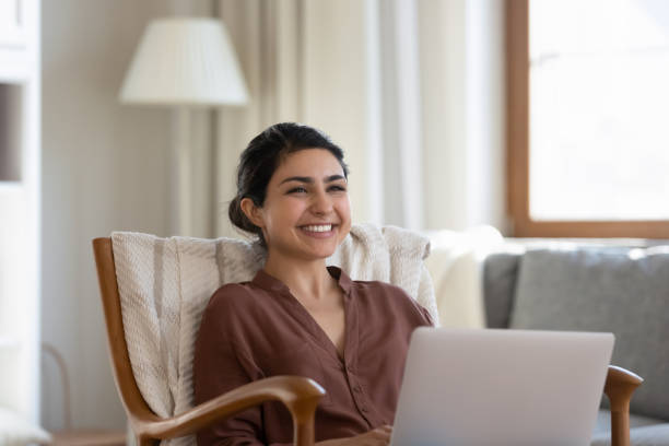 Smiling young indian woman resting in armchair with computer on laps. Distracted from computer work smiling millennial indian ethnicity woman looking in distance, sitting in cozy armchair, feeling excited of getting message with good news, daydreaming resting at home. divergent movie review stock pictures, royalty-free photos & images