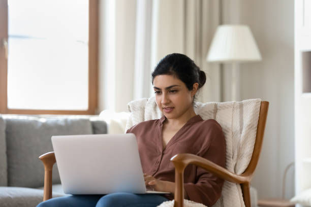 Happy young indian woman typing email on computer. Happy young indian woman typing email on computer, sitting in comfortable armchair, reading media news, web surfing information, planning weekend or vacation trip, playing games or working distantly. divergent movie review stock pictures, royalty-free photos & images