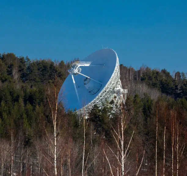 Antenna of the RTF-32 full-rotation precision radio telescope over the forest against the blue sky.  White disk of the radar of radio telescope is with diameter of the main mirror of 32 m and focal length of F = 11.4 m. Svetloye, Leningrad region