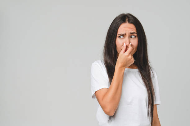 Young disappointed sad caucasian woman in white T-shirt smelling stinky, closing her nose with a hand because of odor stench isolated in grey background stock photo