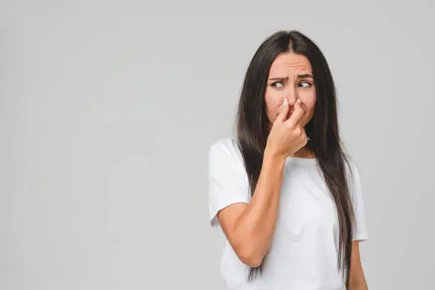 Photo of Young disappointed sad caucasian woman in white T-shirt smelling stinky, closing her nose with a hand because of odor stench isolated in grey background