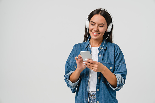 Smiling caucasian young woman listening to the podcast e-book music song singer rock band in headphones earphones, choosing sound track on cellphone isolated in white background