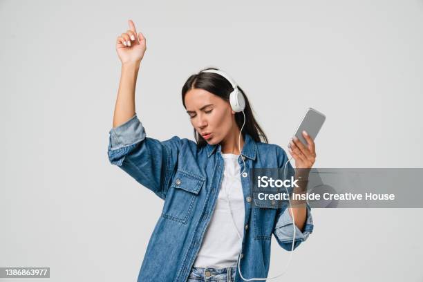 Energetic Caucasian Young Woman Girl Dancing Singing Listening To The Music Podcast Song Singer Sound Track Ebook In Headphones Earphones On Cellphone Isolated In White Background Stock Photo - Download Image Now