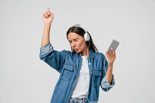 Photo of Energetic caucasian young woman girl dancing singing listening to the music podcast song singer sound track e-book in headphones earphones on cellphone isolated in white background