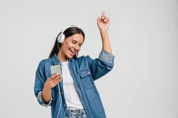 Photo of Dancing singing caucasian young woman using mobile application for listening to music, sound track, playlist, podcast, e-book in headphones isolated in white background