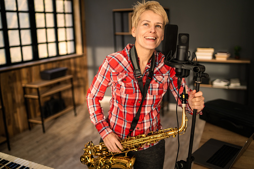 A smiling saxophonist in a recording studio with her saxophone hung from her neck as she has one hand on a microphone stand