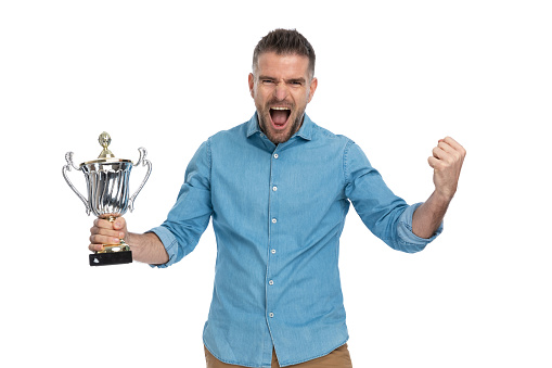 enthusiastic guy in denim shirt holding silver cup, celebrating victory, screaming and being happy for being number one winner on white background