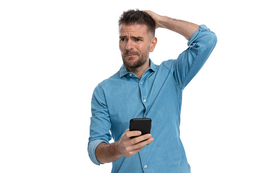 portrait of confused guy in blue jeans shirt scratching head and looking away while holding mobile on white background