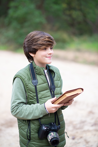 Boy with camera walking in forest. Dark-haired schoolboy in coat holding book. Childhood, nature, leisure concept
