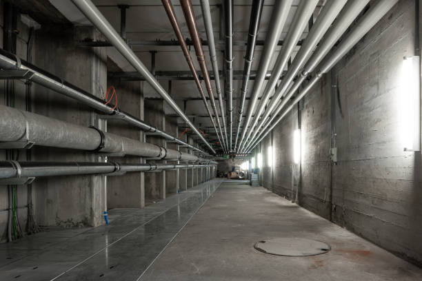 Ceiling mounted industrial pipelines inside underground  building, Long corridor with metal floors, dimly lit, no people stock photo