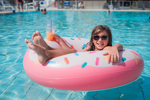 Teenage girl laughing when resting comfortably in a donut shaped swimming float in swimming pool and holding glass of juice