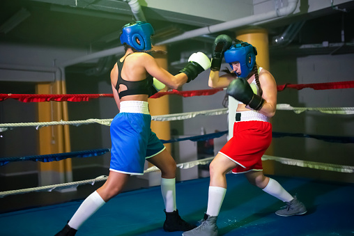 Sporty female boxers boxing in gym. Two trained girls in helmets and gloves practicing sparring on ring, attacking each other making blows, learning to defend. Healthy lifestyle, extreme sport concept