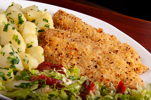 meal with fish fillet or breaded chicken with salad and potatoes cooked on white plate under wooden table