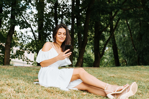 Pretty positive young woman surfs internet, chatting in social media on smartphone while sitting in white summer dress on green grass outdoors. Elegant cute smiling brunette model using mobile phone.
