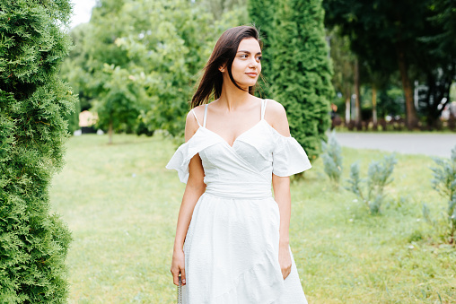 Pretty young brunette woman in white dress with sweet smile looking away in park. Portrait of waist-length elegant model lady standing outdoors on summer day. Fashion and beauty.