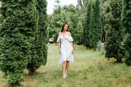 Full length portrait of pretty young brunette woman in white dress with cute smile looking away in park. Elegant lady model with handbag standing outdoors on summer day. Fashion and beauty.