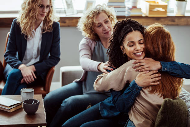 They are having a group therapy session regarding their addiction to recreational drugs. Caring female counselor hugs a female patient during a group therapy session. addict stock pictures, royalty-free photos & images