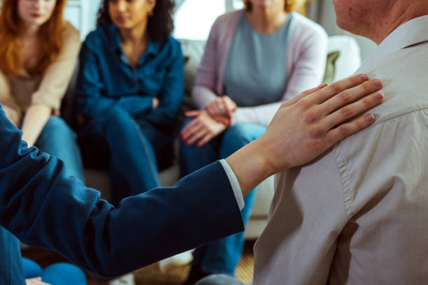 Therapist giving psychological support to a man during therapy session During a support group session, a woman comforts a man; she holds a hand on his shoulder. group therapy stock pictures, royalty-free photos & images