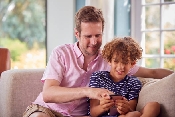 istock Father And Son Sitting On Sofa At Home Playing Game On Mobile Phone Together 1386882546