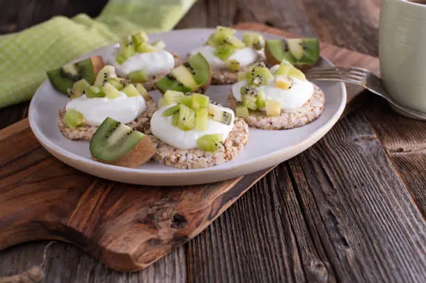 Delicious high protein and healthy breakfast sandwiches. Made with brown rice cakes, topped with skyr and chopped kiwis. Served on a plate on wooden table. Low fat and low calorie fitness food