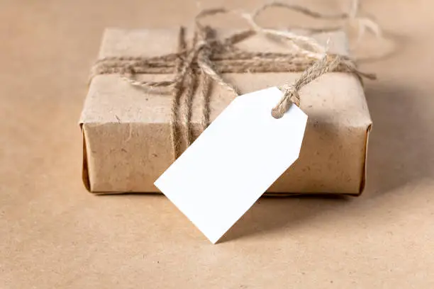 Photo of empty white gift tag mock up on craft paper box on natural beige background.