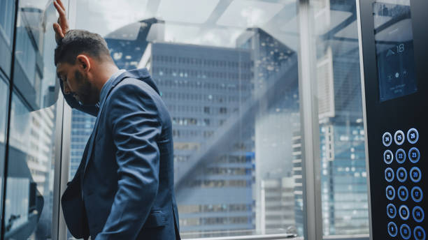 istock Stressed Out Manager Going Down from Office in a Glass Elevator in Modern Business Center. Devastated Specialist Feeling Angry After Unsuccessful Business Meeting, Banging His Head Against a Wall. 1386884135
