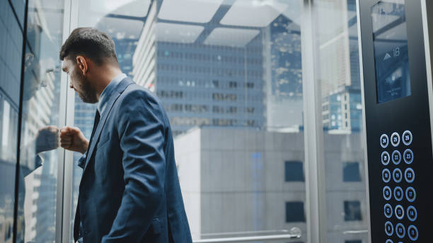 istock Stressed Out Manager Going Down from Office in a Glass Elevator in Modern Business Center. Devastated Specialist Feeling Angry After Unsuccessful Business Meeting, Banging His Knuckle Against a Wall. 1386884144